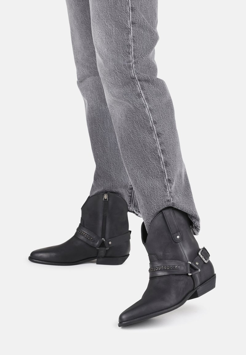 Personne portant un jean gris et des bottines en cuir noir avec fermetures éclair latérales, sangles décoratives et talons bas sur un fond clair.