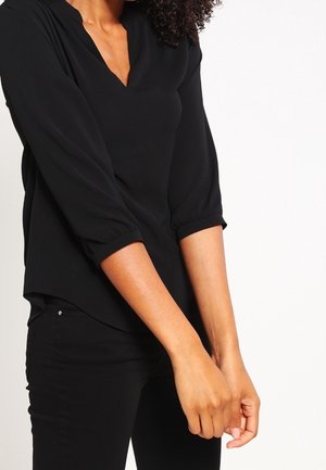 Personne à la peau brun moyen portant un chemisier noir à manches longues et un pantalon noir, les mains jointes devant un fond blanc.