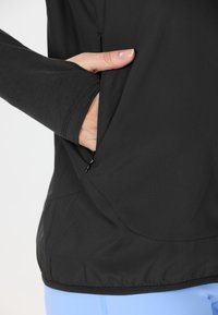 Svart sportjacka med långa ärmar, dragkedjeficka på sidan och rundad nederkant; kombinerad med ljusblå byxor.