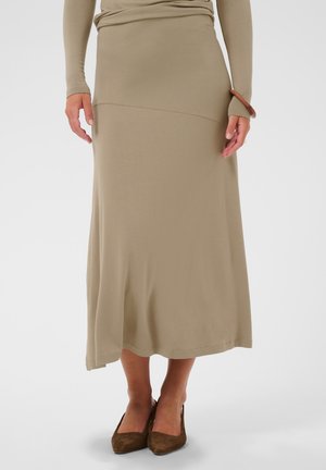 Femme portant une longue jupe beige, un haut beige à manches longues, un bracelet marron et des chaussures marron à bout pointu, debout sur un fond blanc.