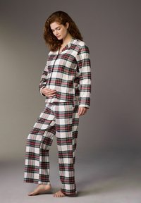 Pregnant woman in red and green plaid pajamas standing barefoot, gently cradling her baby bump against a plain gray background.