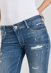 Džínové kalhoty v středním modrém odstínu s opotřebením. Mají pět kapes, zapínání na knoflík a stříbrné kovové akcenty.