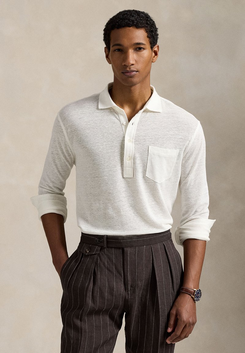 Ανοιχτό μπεζ μακρυμάνικο πόλο με τ σέπη στο στήθος, υφή υφάσματος, και ρολαριστά μανίκια, σε συνδυασμό με σκούρο παντελόνι με λεπτές ρίγες και ρολόι χειρός.