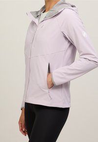 Ljuslila softshelljacka med grå huva, frontdragkedja och sidofickor. Funktionerna inkluderar en mjuk textur och en figurnära design.