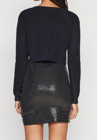 Musta lyhyt neule, jossa poimutettu kuvio, yhdistetty istuvaan, paljeteilla koristeltuun mustaan minihameeseen, joka kimaltelee valon alla.