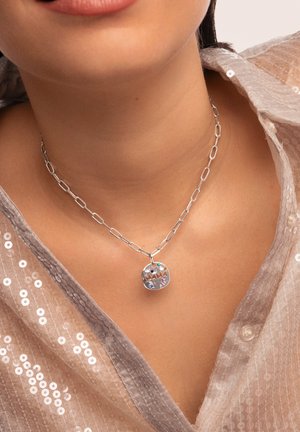 Collier en argent avec un pendentif circulaire arborant le mot "MAMAN" et des accents en pierres précieuses colorées. L'arrière-plan est en tissu texturé.