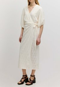 Beige Wickelkleid mit kurzen Puffärmeln, das ein strukturiertes Muster und eine taillierte Bindung hat. Getragen mit braunen Riemchenabsatzschuhen.