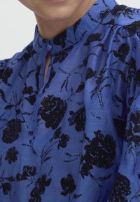 Blusa floreale blu con collo alto, stampa floreale nera, texture sottile e chiusura con bottoni al collo. Maniche leggermente a palloncino.