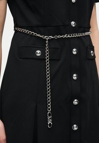 Ασημένια ζώνη αλυσίδα με λογότυπο MK κρέμεται μπροστά πάνω από μαύρο φόρεμα με κουμπιά και τσέπες με καπάκι στη μέση.