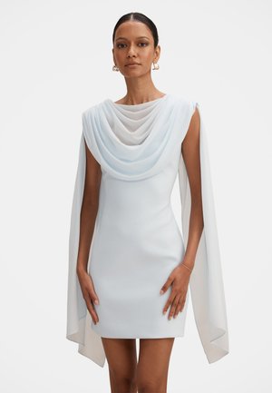 Femme portant une mini-robe blanche sans manches avec un tissu drapé sur les épaules et de longs panneaux fluides en forme de cape descendant le long des bras.