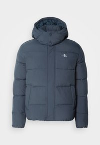 Veste matelassée bleu marine avec un col montant, des boutons-pression et un logo sur le devant. Présente des sections matelassées et des poches latérales. Texture douce.