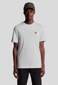 Witte katoenen t-shirt met een klein geel logo op de linkerborst, gecombineerd met een olijfgroen pet. Model staat met de voorkant naar voren.