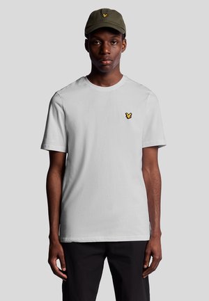 Vit bomulls t-shirt med en liten gul logotyp på vänster bröst, kombinerad med en olivgrön keps. Modellen står vänd mot kameran.
