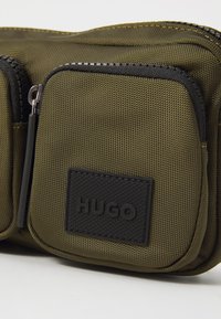 Olivgrünes Nylonetui mit zwei Reißverschlussfächern, schwarzem Zubehör und einem strukturierten schwarzen Logo-Patch mit der Aufschrift "HUGO". Glatte, strapazierfähige Oberfläche.