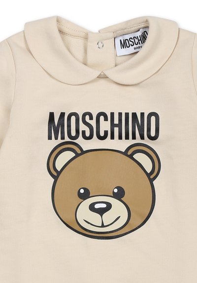 Maglietta per bambino color crema con colletto rotondo, testo nero "MOSCHINO" e grande grafica di un orsetto marrone sulla parte anteriore. Materiale in morbido cotone.