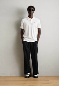 Hvit polo skjorte, svarte vide bukser, svarte sko med hvite detaljer og mørke solbriller. Enkelt design, kontrasterende farger, glatt tekstur.