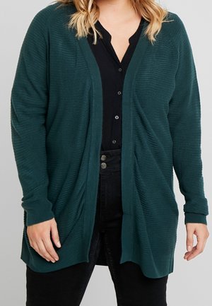 Jadegroene gebreide cardigan met open voorkant, textuurstof, lange mouwen en zijdelingse draping. G gedragen over een zwarte overhemd met knopen en donkere broek.