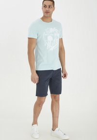 Tricou din bumbac de culoare bleu deschis, cu un imprimeu alb de palmier, asortat cu șorturi navy închise. Adidași albi completează ținuta casual.