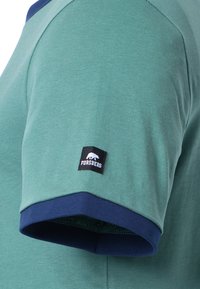 Camisa verde de manga corta con dobladillo azul marino. Presenta una pequeña etiqueta negra con un logo de oso blanco y el texto "FORSBERG" en la manga.
