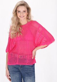 Helles pink gestricktes Sweatshirt mit lockerer Passform, das ein rautenförmiges Netzmuster und kurze Dolmanärmel aufweist. Kombiniert mit Jeans.