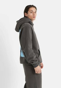 Sudadera gris con un gran estampado gráfico azul en la parte trasera. Presenta un bolsillo frontal, capucha con cordón y puños acanalados. Fondo liso.