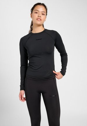 Kvinde med mørkt hår i hestehale iført sort langærmet top og leggings med "hummel"-logos, stående mod en ensfarvet lys baggrund.