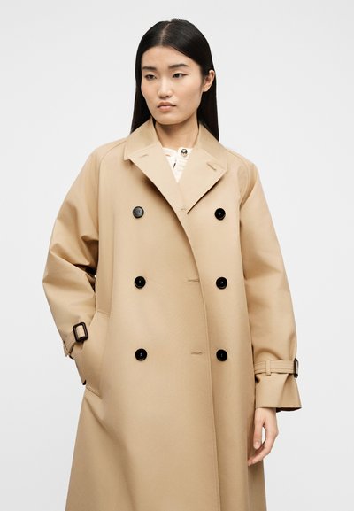 Νεαρή γυναίκα με μακριά σκουρόχρωμα μαλλιά, φοράει ανοιχτό μπεζ καμπαρντίνα με διπλή σειρά μαύρων κουμπιών και μανσέτες με ζώνη, με τα χέρια στις τσέπες.