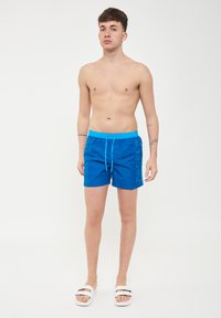Joven de pie descalzo en bañadores azules con texto "SUNDEK" en el lateral y sandalias de doble hebilla blanca contra un fondo blanco.