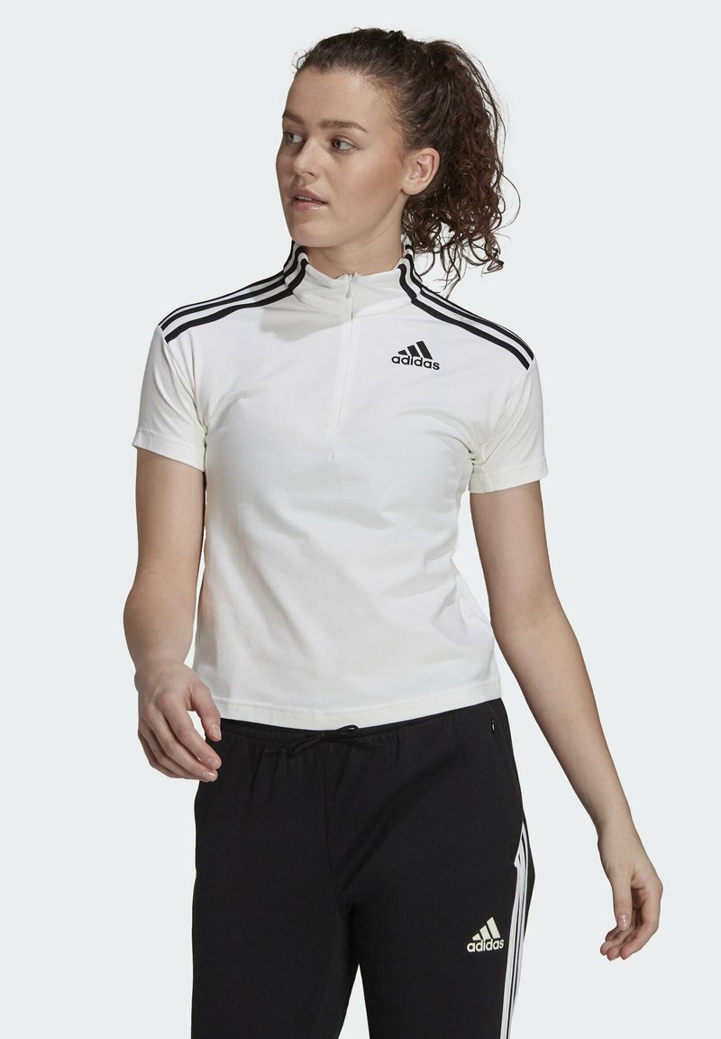 Maglietta bianca a maniche corte con zip, dotata del logo Adidas nero e tre strisce nere sulle spalle, abbinata a pantaloni neri. Texture del tessuto liscia.