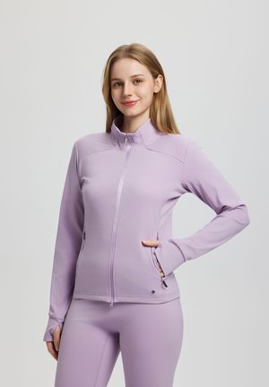 Młoda kobieta z blond włosami ubrana w dopasowaną lawendową kurtkę na zamek i pasujące legginsy, uśmiechająca się, z jedną ręką w kieszeni.