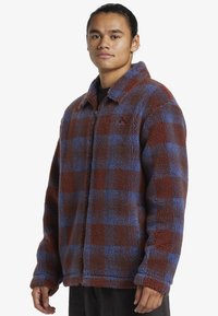 Veste en polaire à carreaux marron et bleu avec col et fermeture éclair. Présente une surface texturée et une coupe décontractée.