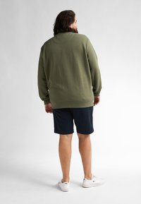 Sudadera texturizada de color verde oliva con cuello redondo, combinada con pantalones cortos azul marino. Modelo usando zapatillas blancas, visto desde la espalda.