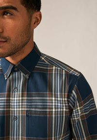 Camisa de botón de color azul oscuro con un patrón de cuadros. Presenta rayas variadas en marrón, crema y verde. Cuello y bolsillo en el pecho destacados.
