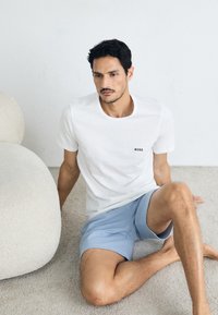 Witte katoenen t-shirt met klein zwart "BOSS" logo, gecombineerd met lichtblauwe shorts. Een gestructureerde, ronde lichtgekleurde bank is in de buurt.
