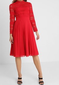 Röd långärmad klänning med en texturerad spetskjol, plisserad kjol och en figurnära midja. Matchad med svarta spänneklackar.