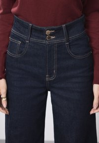 Kõrge vöökohaga tumedat sinist teksapüksid kuldsete detailide, kahe nupuga kinnituse ja kontrastõmblustega. Omavad külg- ja tagataskuid.