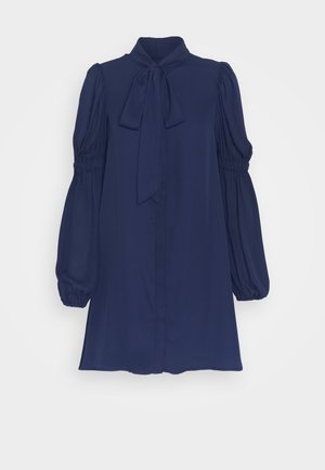 Blouse bleu marine en tissu léger, présentant un col montant, un détail noué et des manches bouffantes avec des poignets élastiques.