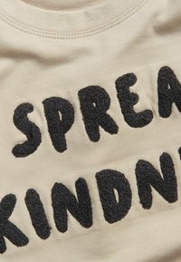 Beige bomullströja med texturerad, fluffig svart lettering som tydligt säger "SPREAD KINDNESS" över framsidan.