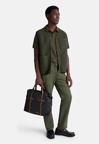 Muž oblečený v olivově zeleném polo tričku, odpovídajících kalhotách a vestě, držící černou a hnědou koženou tašku s rukojeťmi, stojící na bílém pozadí.