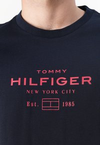 Tamsiai mėlynas medvilninis marškinėlis su raudona grafine užrašu "TOMMY HILFIGER NEW YORK CITY Est. 1985" ir kvadratiniu logotipo akcentu. Klasikinis apvalus kaklo iškirpimas.