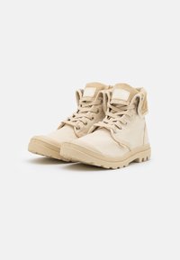 Las botas altas beige cuentan con una parte superior de lona, cordones y una suela de goma texturizada. Parche del logo destacado en la lengüeta.