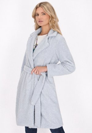 Donna che indossa un cappotto chiaro grigio peloso con cintura sopra una maglietta bianca e pantaloni scuri, posa con la mano che tiene la cintura del cappotto.