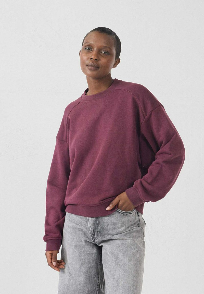 Felpa bordeaux con collo rotondo e maniche raglan, realizzata in tessuto morbido. Abbinata a jeans grigi chiari con una vestibilità rilassata.