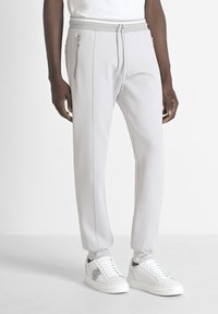 Man in lichte grijze joggers met ritssluitingen aan de zijkant en witte sneakers met zwart-wit embleemdetail.
