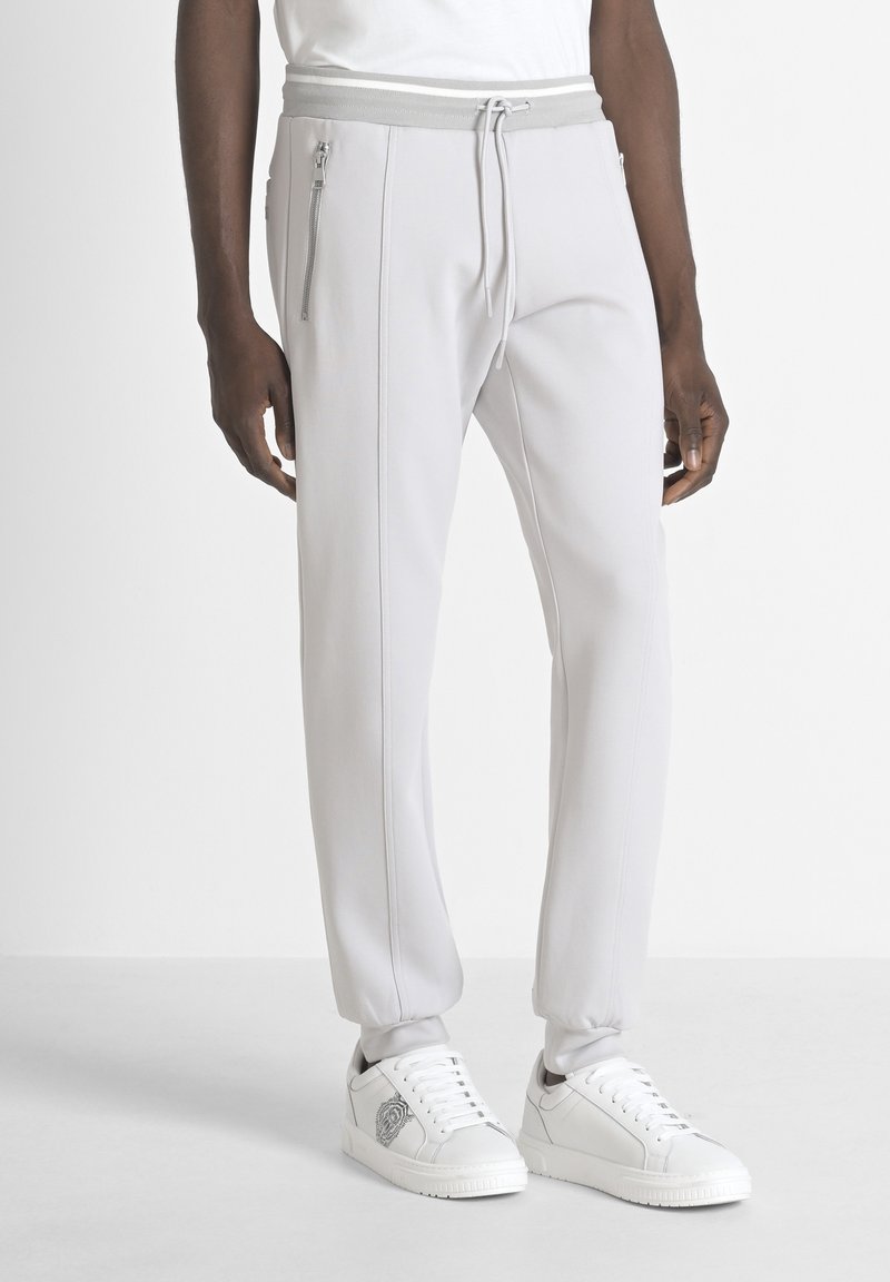 Man in lichte grijze joggers met ritssluitingen aan de zijkant en witte sneakers met zwart-wit embleemdetail.