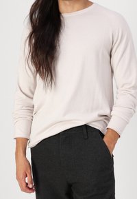 Helle beige Langarmhemd mit rundem Ausschnitt und gerippten Bündchen, kombiniert mit dunkelgrauen Hosen mit Seitentasche. Glatte Textur.