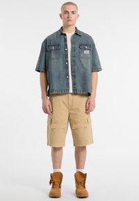 Chemise en denim à manches courtes, associée à un short cargo beige et des bottes de travail brunes. Comprend deux poches poitrine et une coupe décontractée.