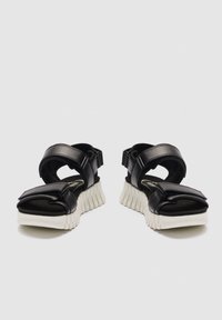 Černé sandály s otevřenou špičkou, silnou bílou rýhovanou podrážkou a nastavitelnými pásky přes prsty a kotníky na světlém pozadí.