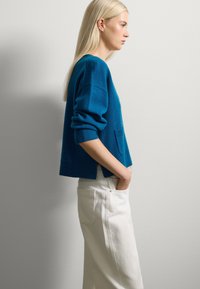 Suéter de lana turquesa con mangas tres cuartos, puños acanalados y un bolsillo, combinado con pantalones anchos blancos, frente a un fondo neutral.
