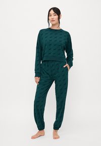 Nainen seisoo paljain jaloin päällä tummanvihreä yhteensopiva pitkähiha toppi ja jogger-housut, joissa on hienovarainen toistuva logo-kuvio.